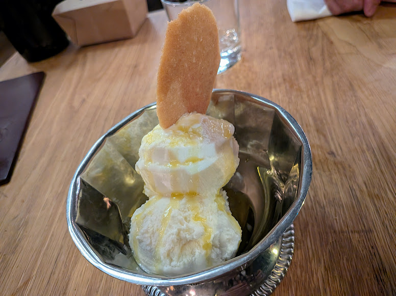 Two scoops of vanilla ice cream topped with a thin biscuit and drizzled with syrup, served in a silver dish on a wooden table.