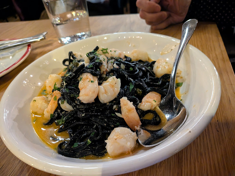 A plate of black squid ink pasta topped with shrimp and herbs, served with a large spoon on a wooden table.