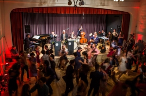 A band performs on stage with various instruments while people dance energetically on the wooden floor in front of them.