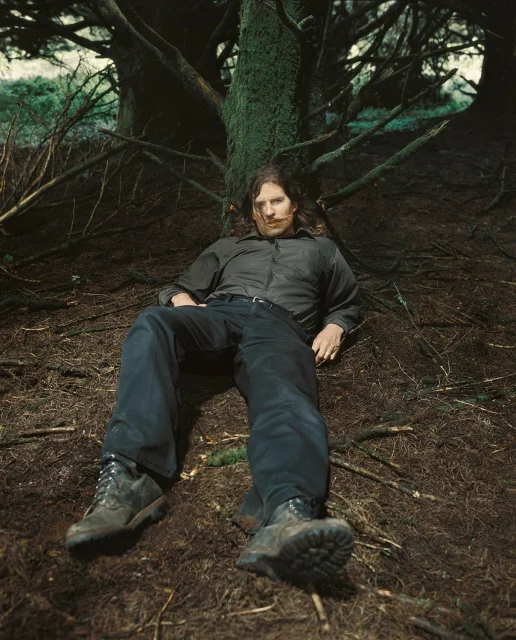 A man with long hair, wearing dark outdoor clothing and boots, sits slouched against a tree trunk in a forest with dense branches and uneven ground.