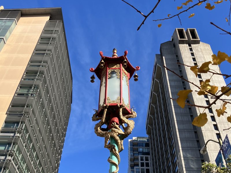 A traditional Chinese-style street lantern with a dragon design stands between two modern buildings under a clear blue sky, with yellow leaves framing the top right corner.