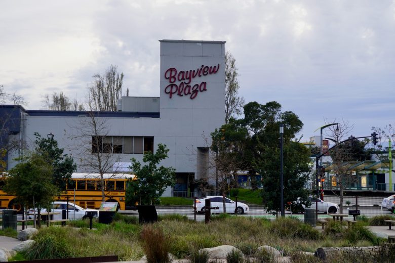 Bayview Plaza building with a large sign, visible trees and plants in the foreground, yellow school bus, and cars on the street.