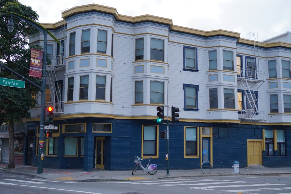 A three-story blue and white corner building with yellow trim, large windows, and a bike parked on the sidewalk near a traffic light and street signs.