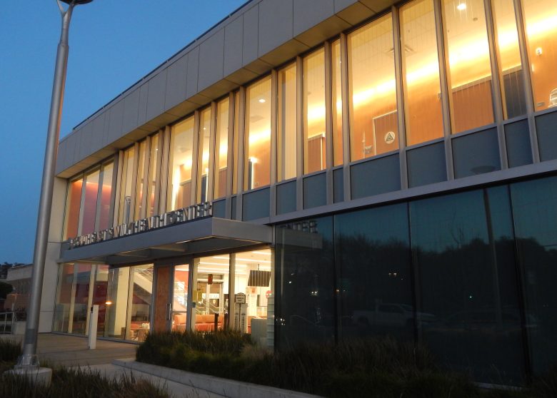 A modern two-story building with large glass windows and illuminated interior lights, photographed at dusk.