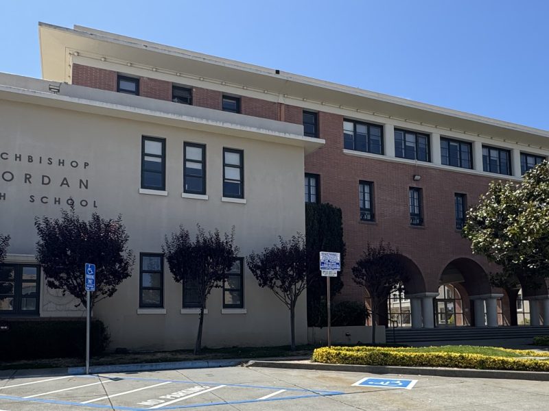 A three-story building with "Archbishop Riordan High School" on the facade, featuring accessible parking spaces and trimmed trees in front.
