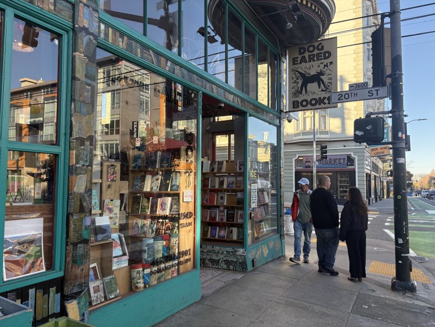 Street view of Dog Eared Books bookstore at a corner, with book displays in the window and three people walking by on the sidewalk.