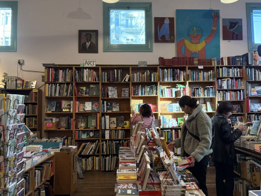 Three people browse books in a well-lit bookstore with shelves filled with books, two artworks and three windows visible on the back wall.