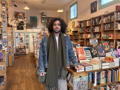 A person with curly hair stands in a bookstore, resting a hand on a display of books. Shelves filled with books and eclectic decor are visible in the background.