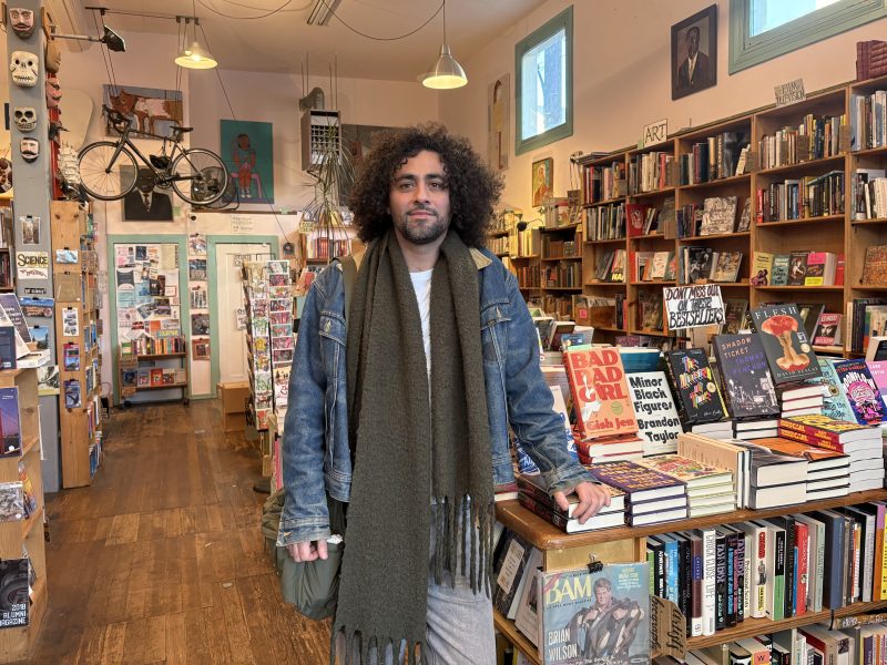A person with curly hair stands in a bookstore, resting a hand on a display of books. Shelves filled with books and eclectic decor are visible in the background.