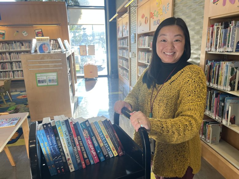 A woman stands in a library next to a cart filled with books, smiling at the camera. Bookshelves and children's area are visible in the background.