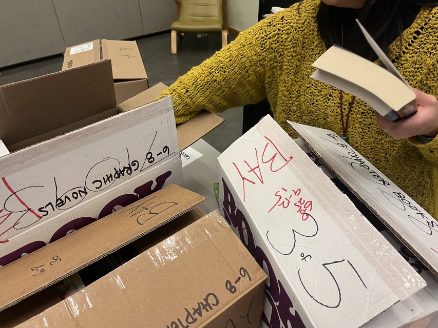 Person in a yellow sweater organizing books into cardboard boxes labeled with age groups and genres, holding a paperback book in one hand.