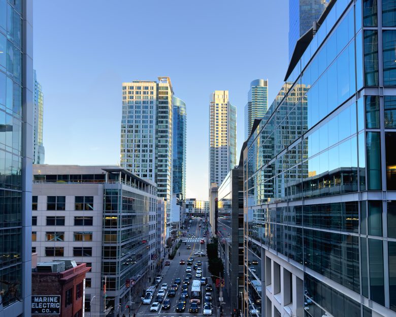 A city street lined with modern glass buildings and high-rises, with cars and light traffic visible during daylight.