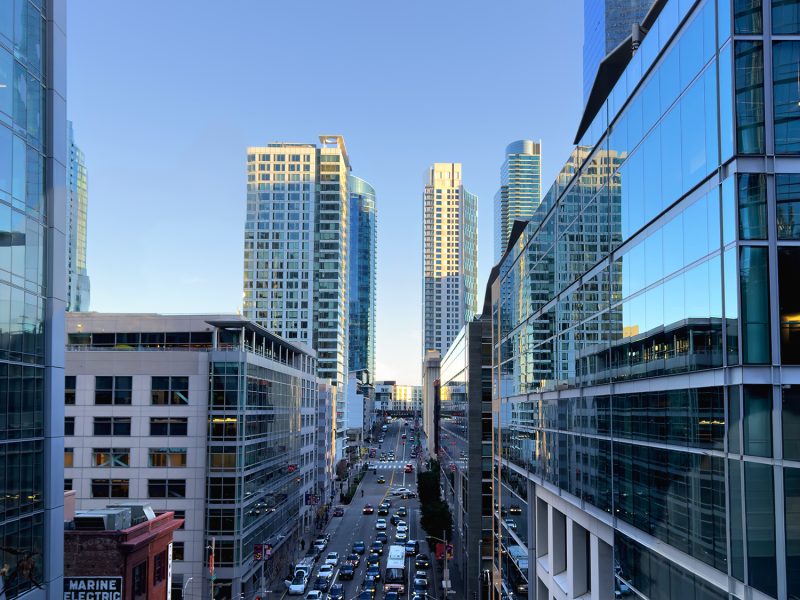 A city street lined with modern glass buildings and high-rises, with cars and light traffic visible during daylight.