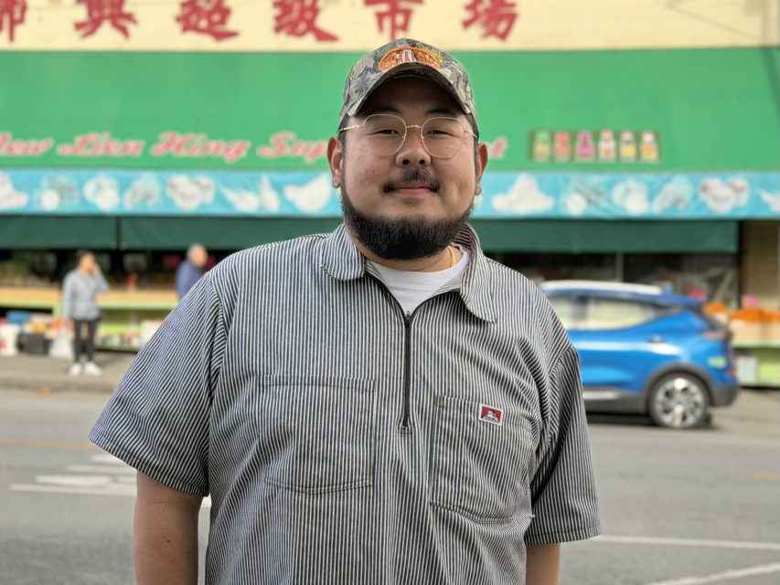 A man wearing glasses, a striped shirt, and a camouflage cap stands on a city street in front of a green awning and storefront.