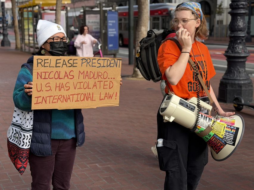 Two people stand on a city sidewalk; one holds a sign about Nicolas Maduro and U.S. violations, the other holds a megaphone covered in stickers.