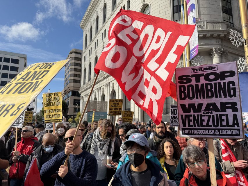 A crowd of protesters holds signs reading “PEOPLE POWER,” “STOP BOMBING VENEZUELA,” and “FOR JOBS AND EDUCATION” at a demonstration in an urban area.