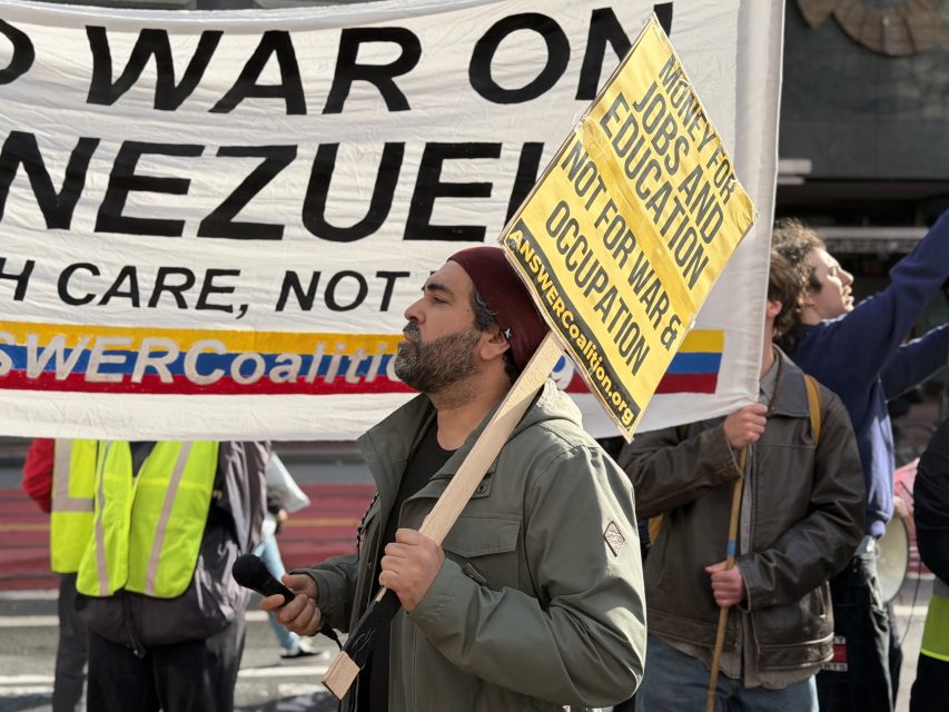 A man holds a yellow protest sign reading "Money for jobs and education, not for war and occupation" at a demonstration against war in Venezuela. Other protestors and banners are visible behind him.