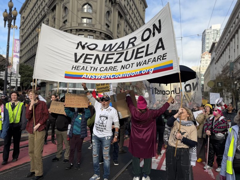 A group of protesters in a city street hold a large banner reading “No War on Venezuela. Health Care, Not Warfare. ANSWERcoalition.org.” Other signs and people are visible around them.