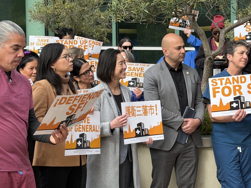 A diverse group of people stand outdoors holding “Stand Up for SF General” and “Save SF General” signs in multiple languages at a public demonstration.