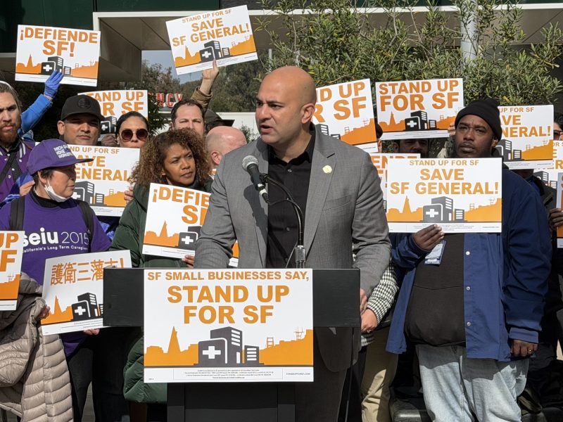 A man speaks at a podium during a rally, surrounded by people holding signs that read “Stand up for SF” and “Save SF General!” in support of San Francisco small businesses and hospitals.