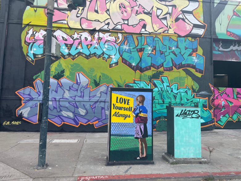 A utility box on a sidewalk features art of a person holding flowers with the text "LOVE Yourself Always"; colorful graffiti covers the wall in the background.