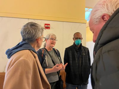 Four adults stand talking indoors; one woman gestures with her hands, another has short gray hair and glasses, a man wears a mask, and another man faces away from the camera.