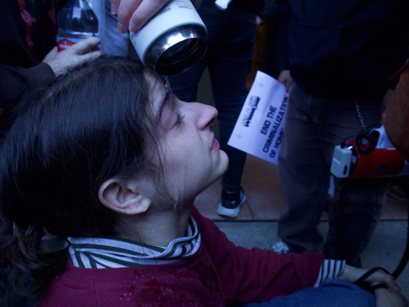 A person pours liquid from a bottle onto another person's face, who appears to be sitting on the ground with eyes closed. Others stand nearby, one holding a sign.