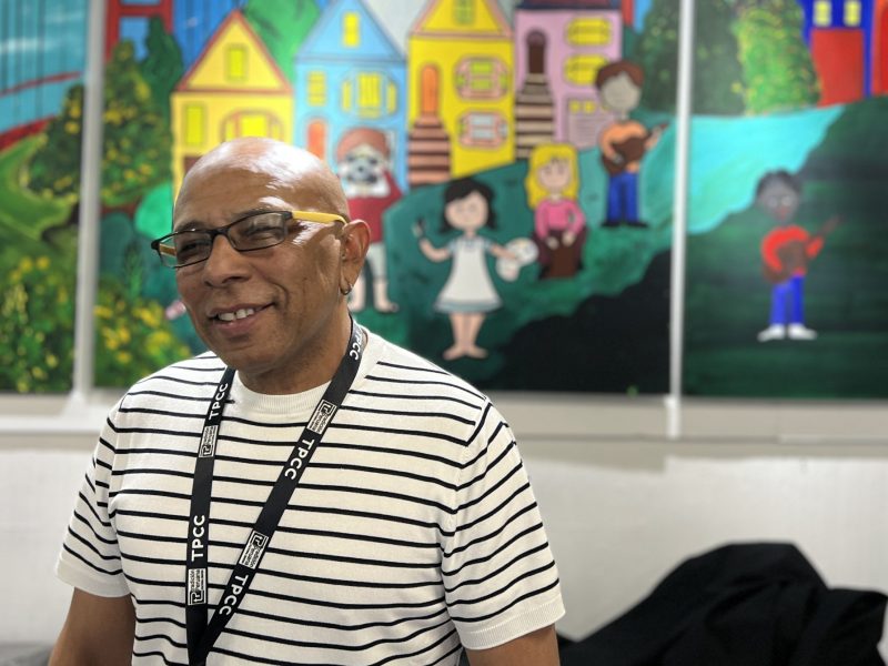 A man in a striped shirt stands smiling in front of a colorful mural depicting houses and people in front of the Golden Gate Bridge.