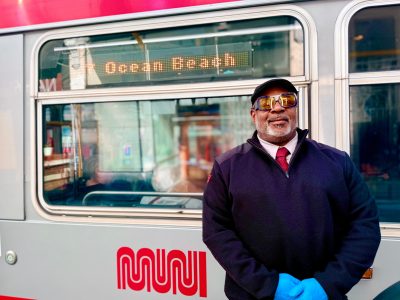 A transit worker in uniform and blue gloves stands in front of a Muni light rail vehicle displaying "Ocean Beach" as its destination.