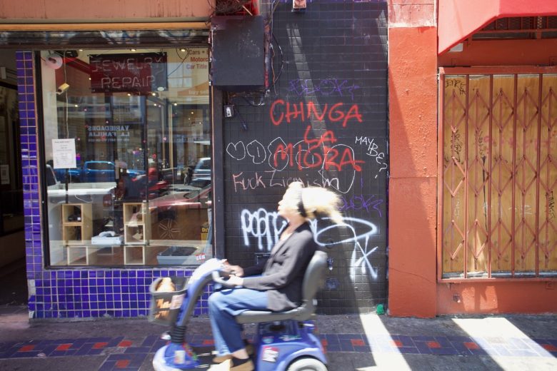 A person rides a mobility scooter past a wall with graffiti and a boarded-up storefront in an urban setting.