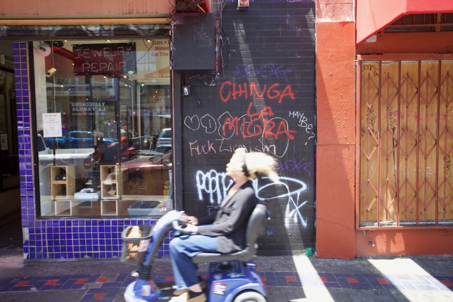 A person in a mobility scooter passes by a graffiti-covered wall and storefront in an urban area during daytime.
