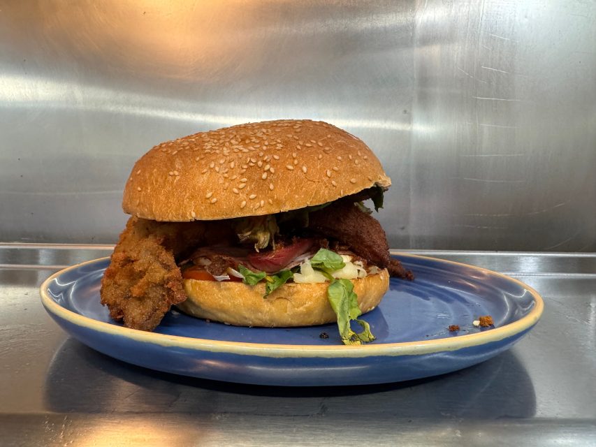 A fried chicken sandwich with lettuce and other toppings on a sesame seed bun, served on a blue plate against a metal background.