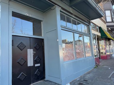 A light blue storefront with large windows and double black doors at 3355; nearby buildings have awnings and graffiti is visible on the window.
