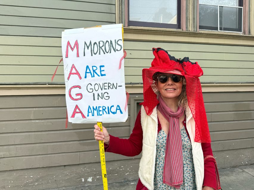 A woman wearing sunglasses and a red hat holds a sign reading "MAGA Morons Are Governing America" in front of a beige wooden building during an ICE walkout.