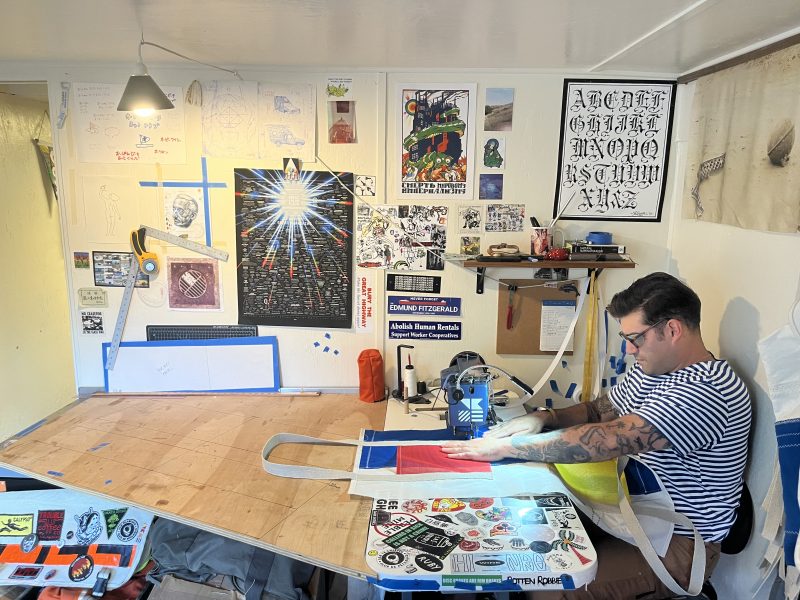 A man wearing glasses and a striped shirt works with fabric on a large table in an art studio filled with posters and tools.