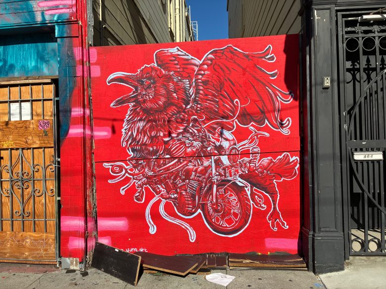 A mural of a large red raven with mechanical and motorcycle elements is painted on a red wall between two buildings.