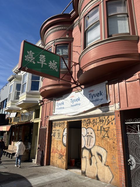 A red building with bay windows and a green sign with Chinese characters; ground level has Tyvek wrap and graffiti, with a partially open door and people walking by.