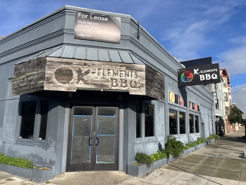 Gray building with boarded windows and "K Elements BBQ" signs; a "For Lease" sign is visible on the roof, indicating the space is available for rent.