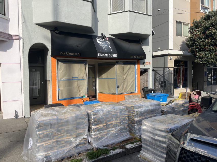 Stacks of construction materials are piled on the sidewalk in front of a closed restaurant with "Umami Sushi" signage and windows covered in brown paper.