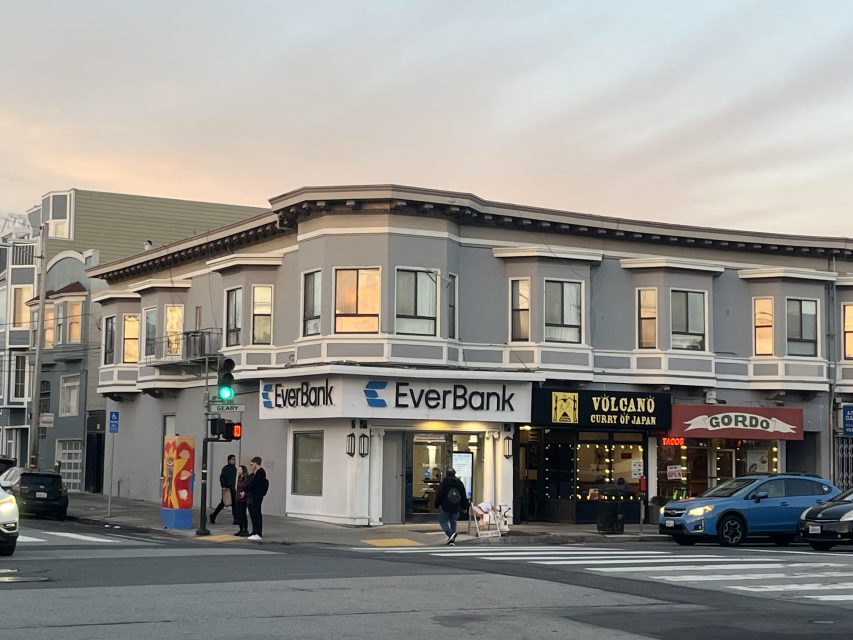 A gray two-story corner building houses EverBank, Volcano restaurant, and Gordo eatery. People stand on the sidewalk and cars are parked along the street at sunset.