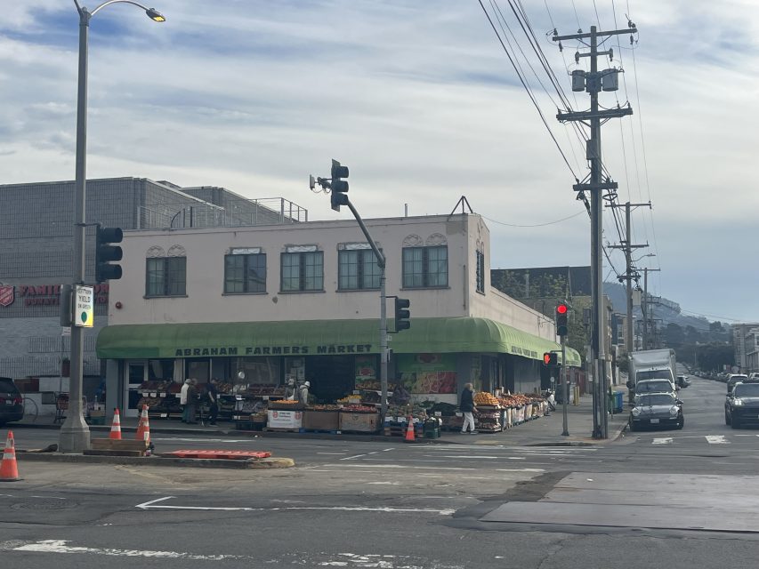 A two-story building with a green awning labeled "Abraham Farmers Market" stands on a street corner with fruit and vegetable displays outside.