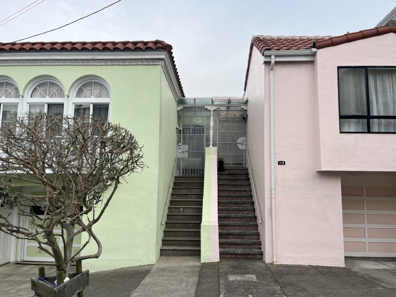 Two pastel-colored houses, one green and one pink, separated by a staircase leading to a gated entry; a leafless tree is in front of the green house.