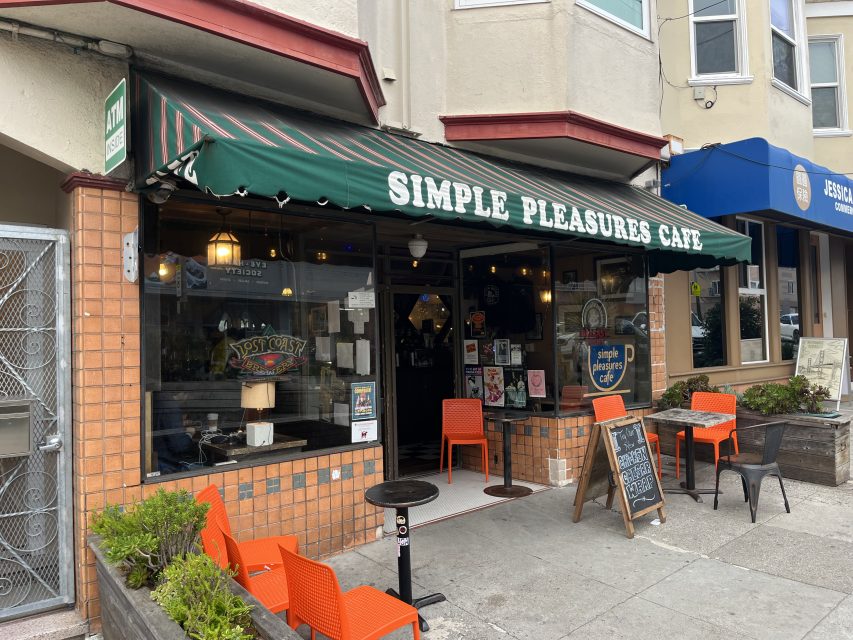 Exterior of Simple Pleasures Cafe with green striped awning, orange chairs and tables outside, chalkboard menu, and plants along the sidewalk.