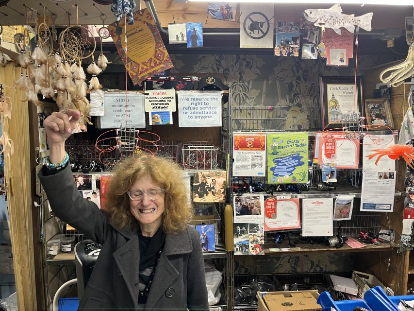 A woman with curly hair smiles and raises her hand inside a shop filled with various hanging items, signs, and colorful decorations behind the counter.
