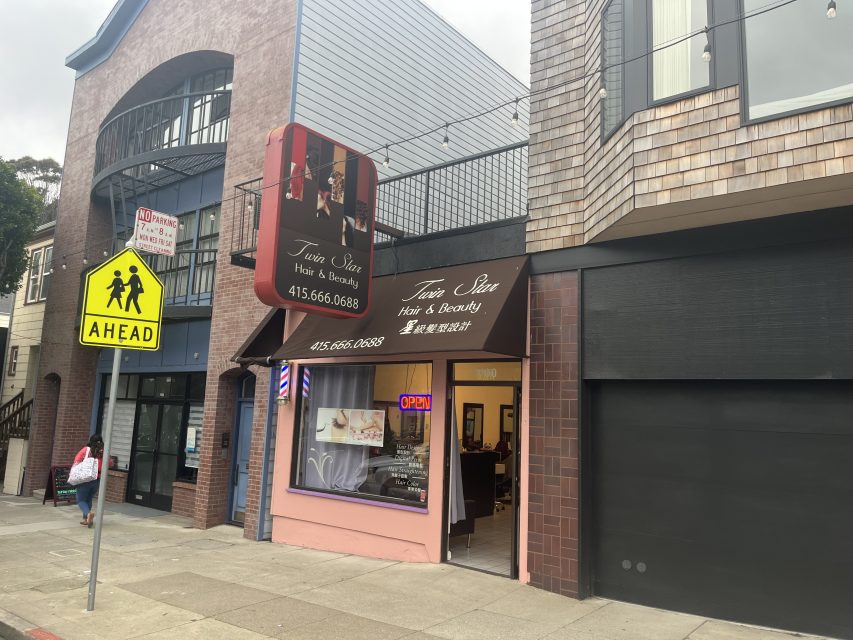 Storefront of Twin Star Hair & Beauty with an "OPEN" sign, a sidewalk, a yellow pedestrian crossing sign, and nearby buildings visible.