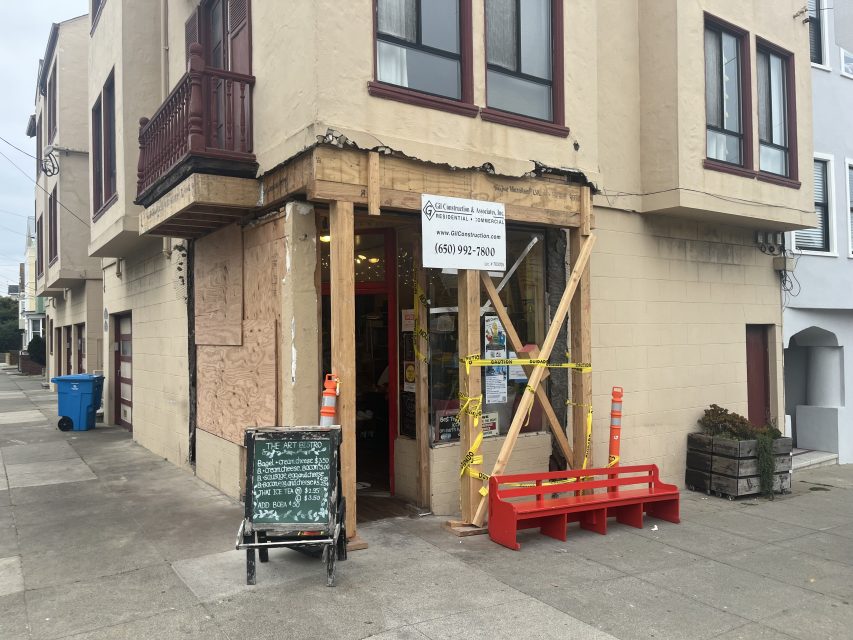 A corner store entrance under construction with wooden supports, yellow caution tape, orange cones, and a sandwich board on the sidewalk.