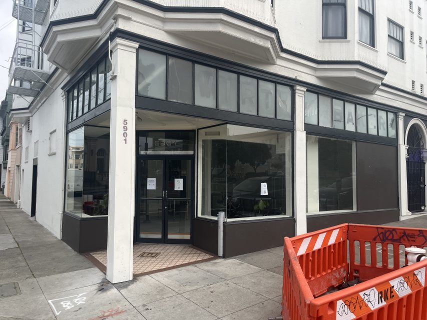 A vacant storefront on a street corner with large glass windows, tiled entryway, and a numbered address “5901”; an orange barricade is visible on the sidewalk in front.