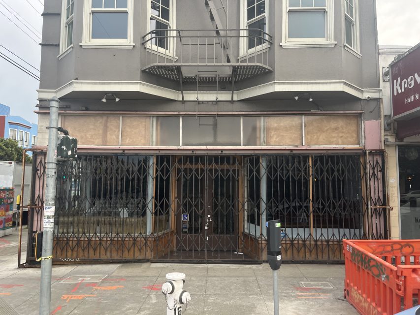 A closed storefront with metal security gates pulled shut, boarded sign area above, and a fire escape on the facade. A parking meter and fire hydrant are visible on the sidewalk.