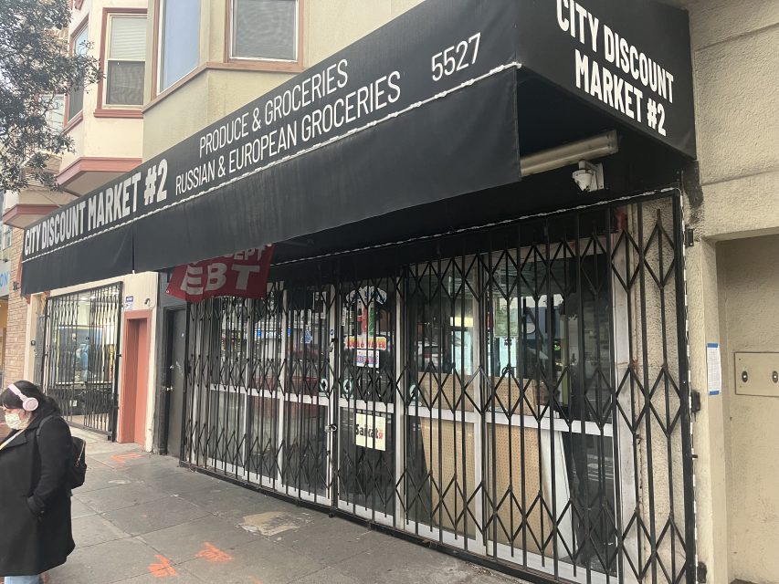 A closed storefront with a black awning reading "City Discount Market #2" and "Produce & Groceries, Russian & European Groceries." A person wearing a mask walks by.