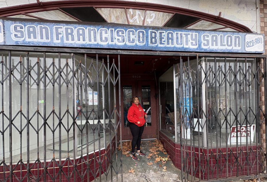 A woman in a red hoodie stands at the entrance of the closed San Francisco Beauty Salon, with security gates partially open and fallen leaves on the ground.